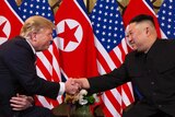 Donald Trump and Kim Jong-un shake hands while they sit in chairs in front North Korean and United States flags.