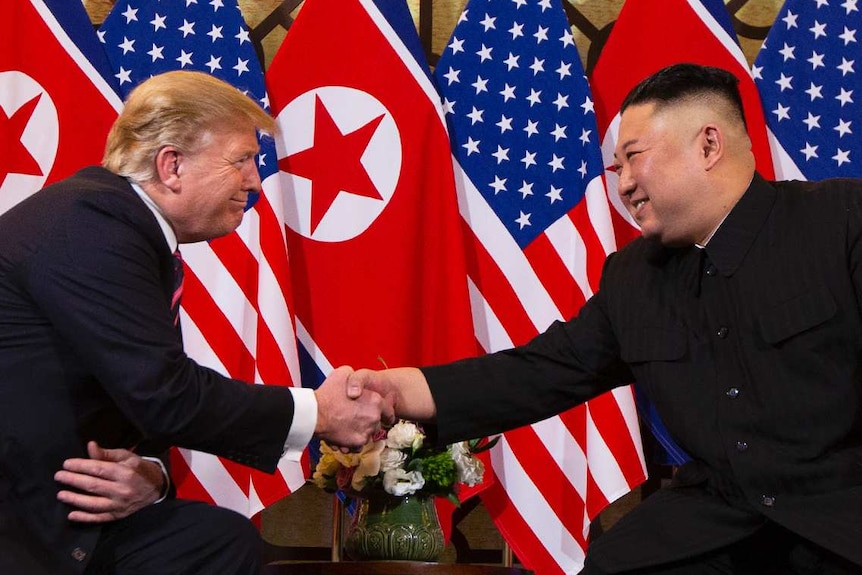 Donald Trump and Kim Jong-un shake hands while they sit in chairs in front North Korean and United States flags.