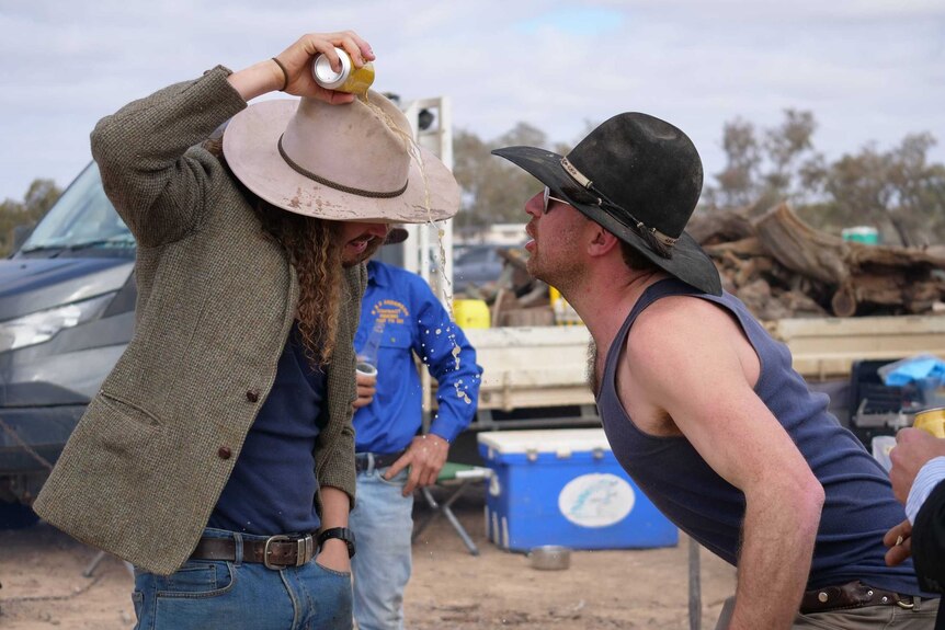 Two men wearing Akubra-style hats face each other as one pours beer over his hat.