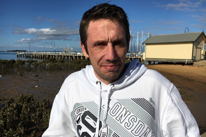 A man wearing a white hoodie looks towards the camera with a beach  mudflat and boat masts in the background.