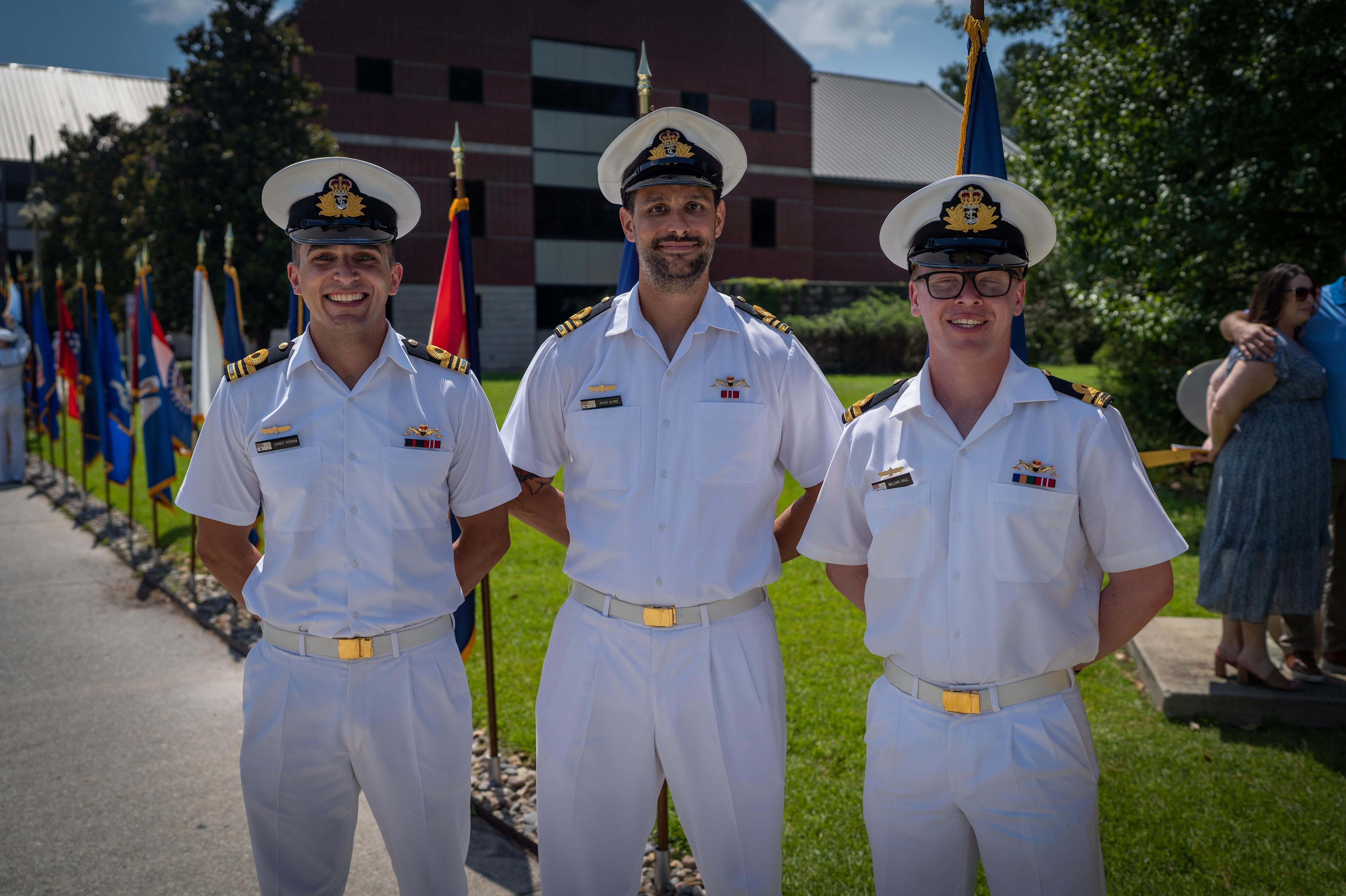 Three men in white naval uniform stand side by side, hands behind their backs, outside on bright day