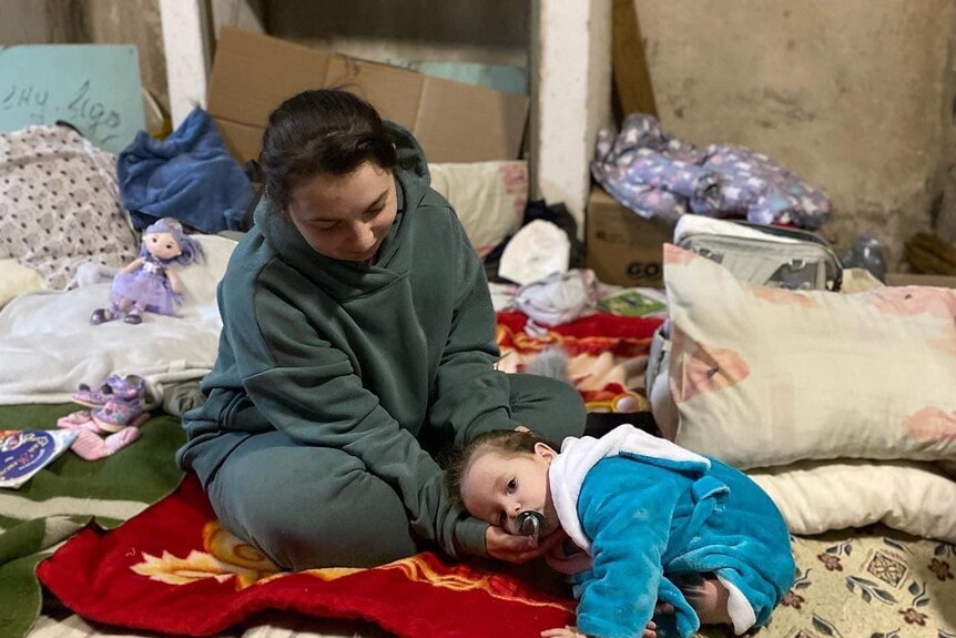 A woman sits on cushions on the floor, with a small baby laying next to her. They are in a basement.
