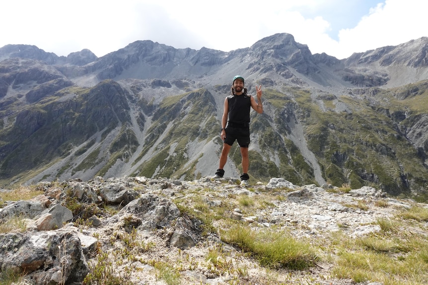 Josh Storey gives the peace sign while standing with large mountains behind him, wearing a black singlet and shorts.