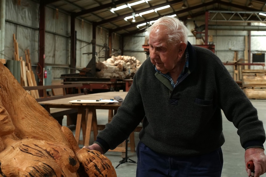 A man with white hair touches a carved piece of wood in a sawmill.