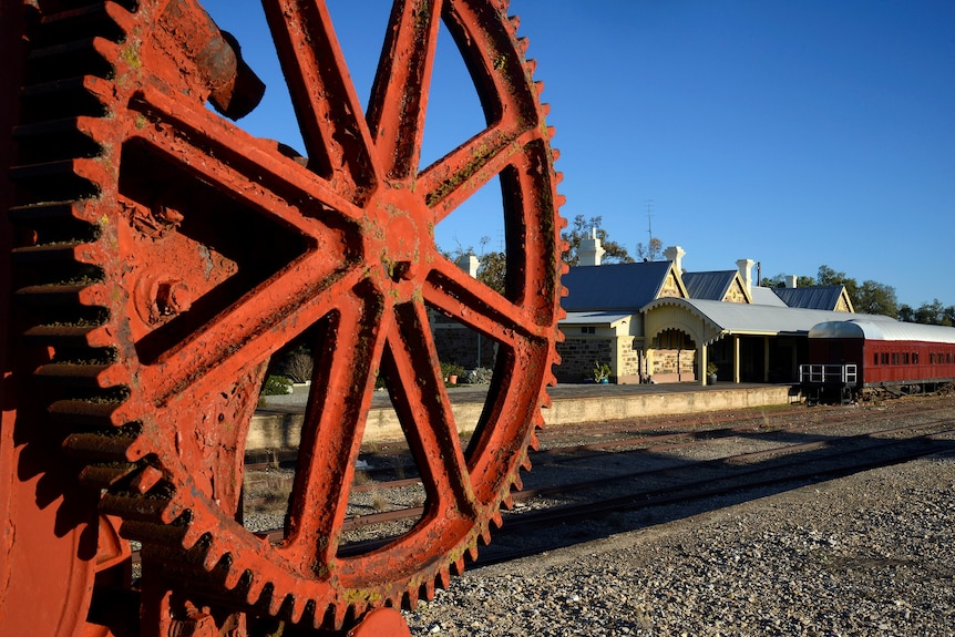 A giant cogged wheel in the foreground, with the station and a train in the background