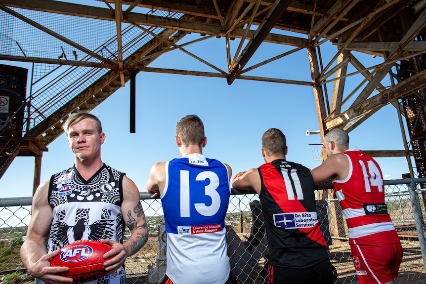 A man in a jersey holding an AFL football standing next to three other men wearing different coloured jerseys facing away.