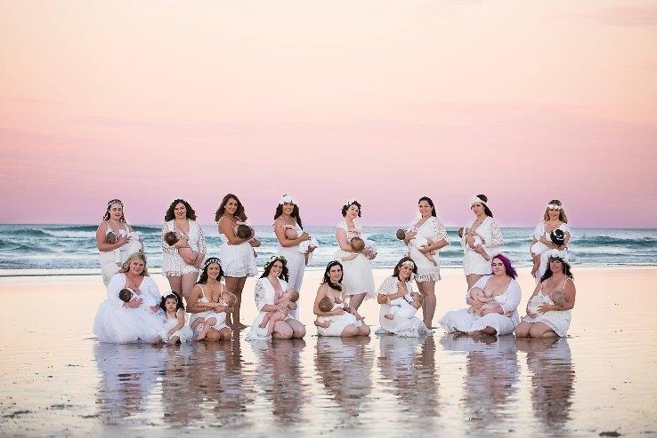 Fifteen women breastfeeding their babies on a beach at sunset.