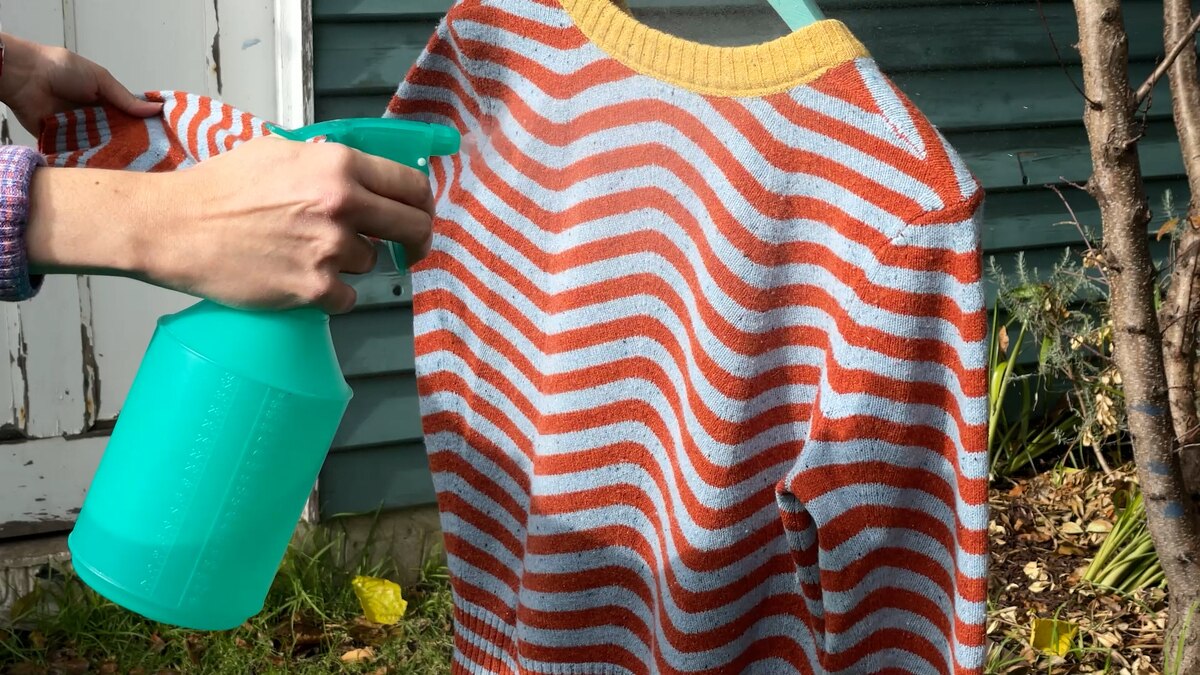 Close up picture of woman spraying a stripy jumper with vodka water, the spray bottle left of the screen in front of garden shed