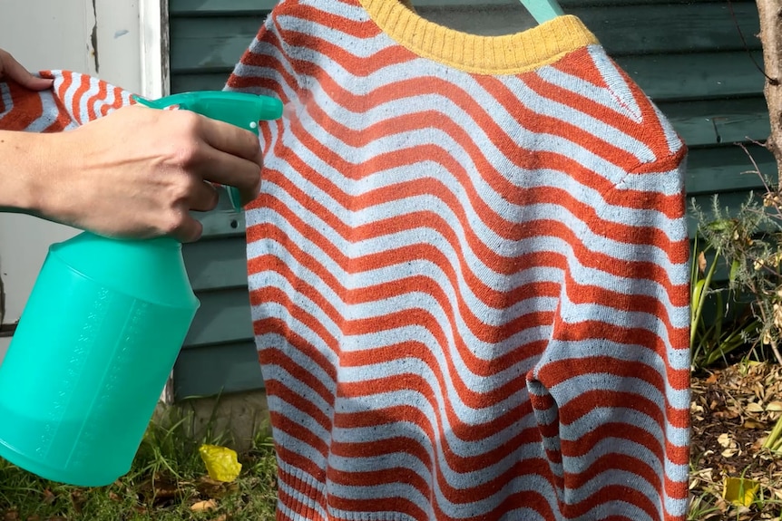 Close up picture of woman spraying a stripy jumper with vodka water, the spray bottle left of the screen in front of garden shed