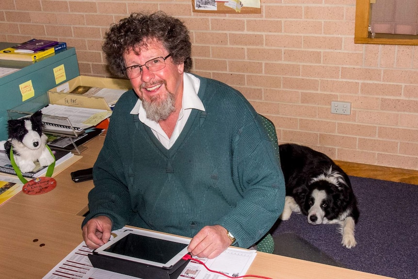 A priest in ordinary clothes sits at a desk in an office with a border collie dog lying on the floor beside him