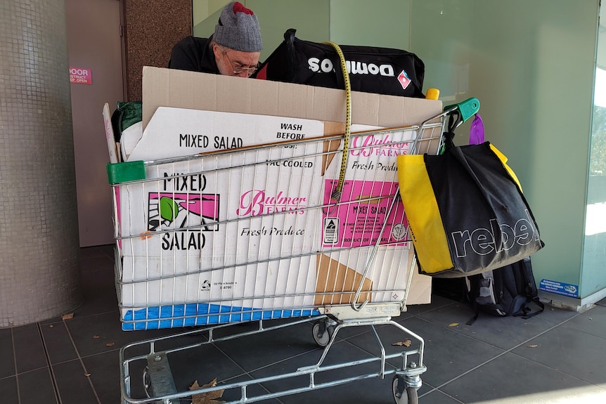 A man with a beanie standing behind a trolley full of cardboard and other bits and pieces. He is reaching inside it.