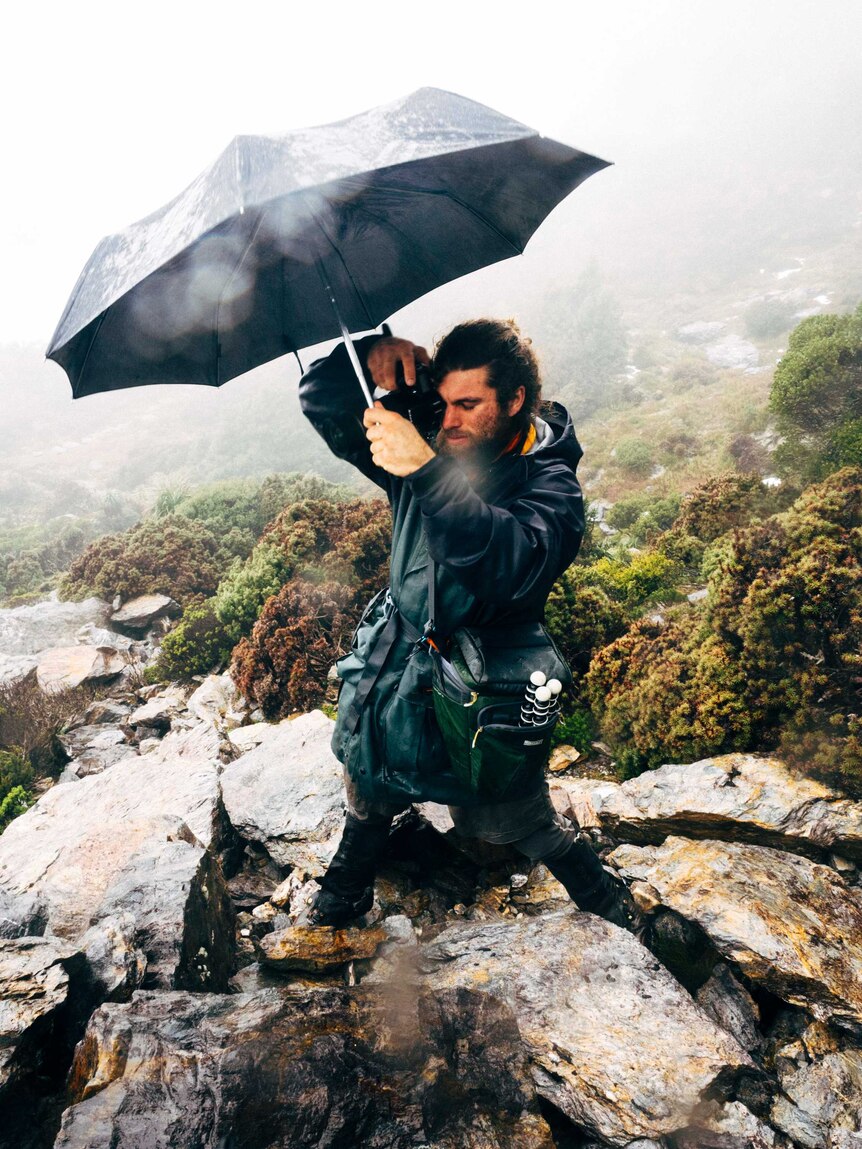 Dan Haley holding camera in rain under umbrella.