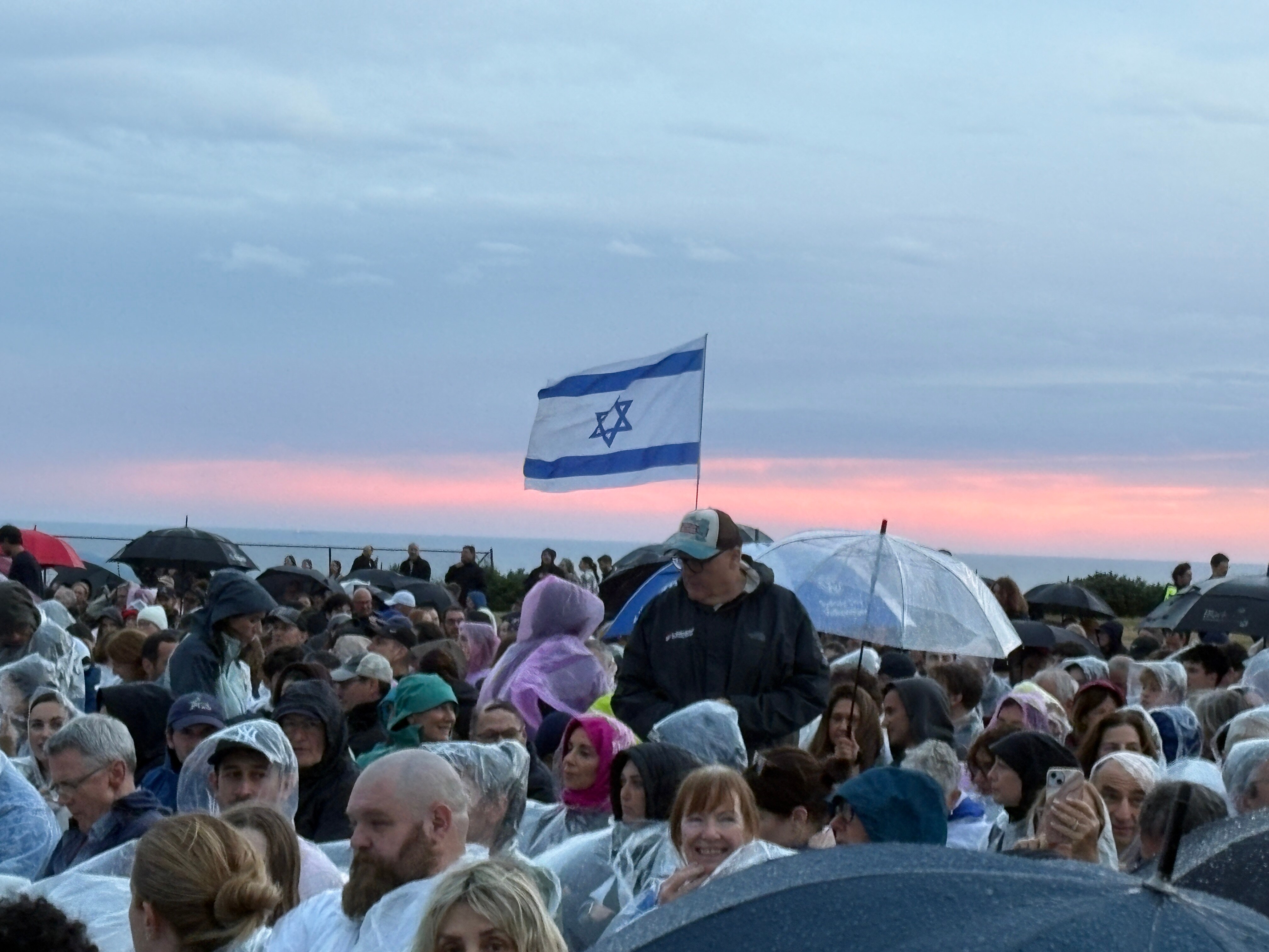People gathered at dusk, blue and white Israeli flag flying among crowd