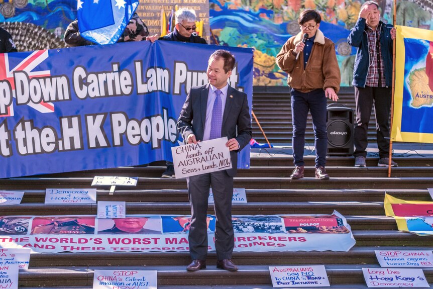A man in a navy suit holds a placard that reads 'China hands off Australian seas + lands now' surrounded by similar banners.