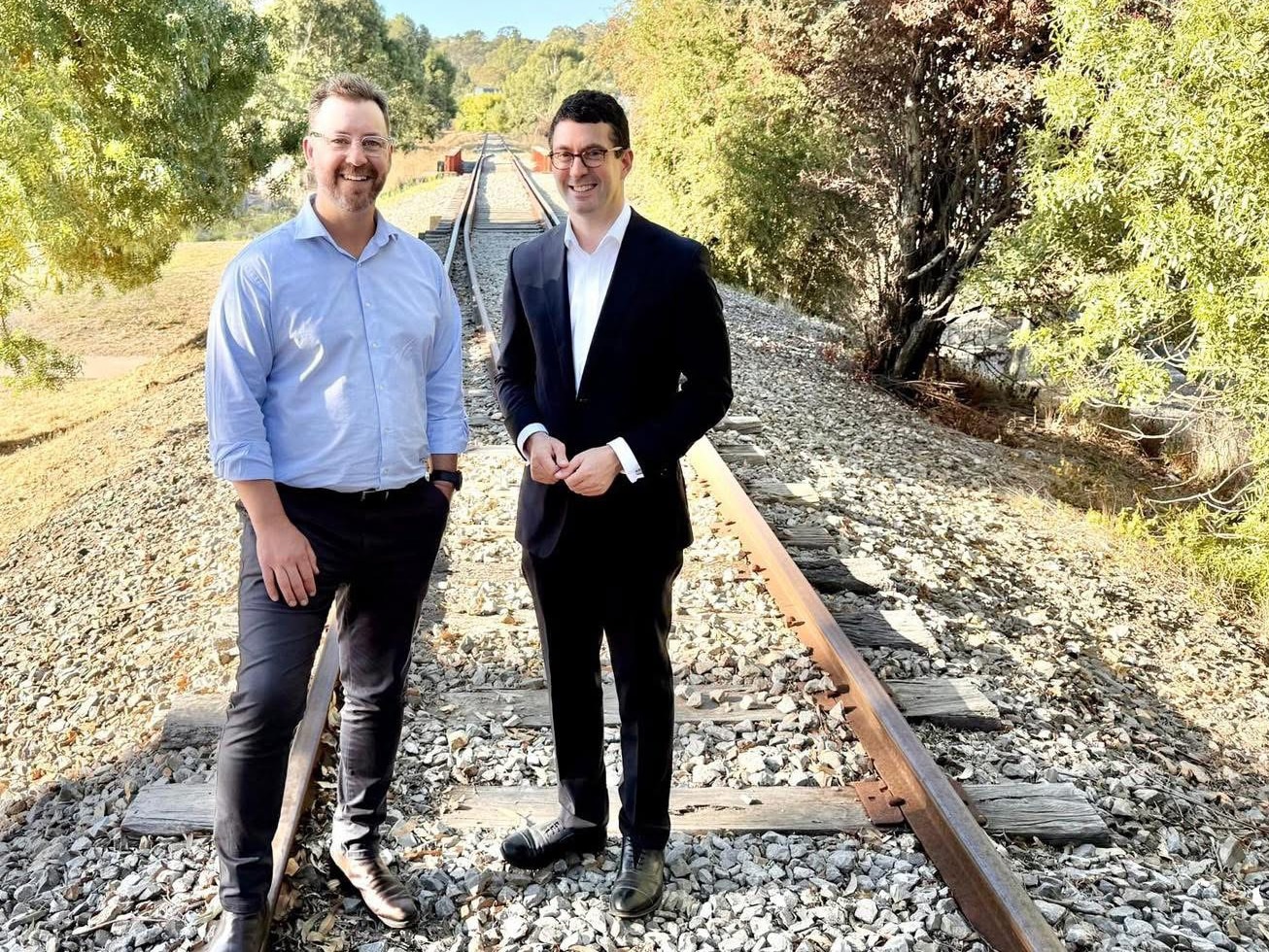 Matt con una camisa azul, jeans negros y Dan con un traje negro, camisa blanca, parados en una vía de ferrocarril en el monte.