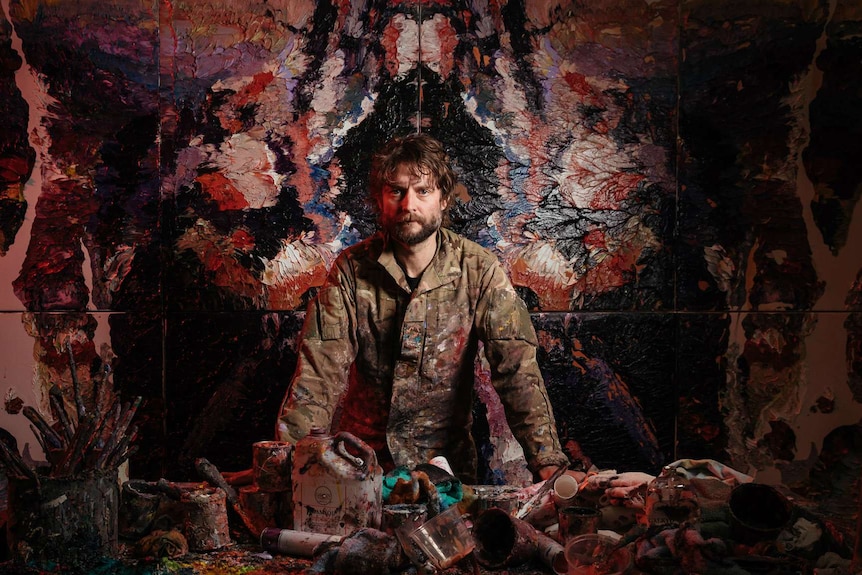 A young man covered in paint stands in front of an enormous painting