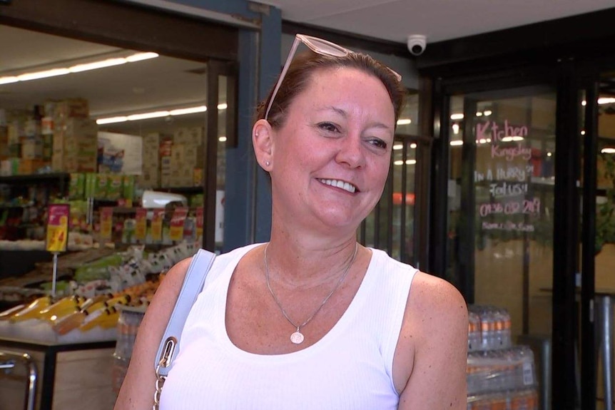 A close up of a woman with sunglasses on her head wearing a white singlet top in front of a shop.
