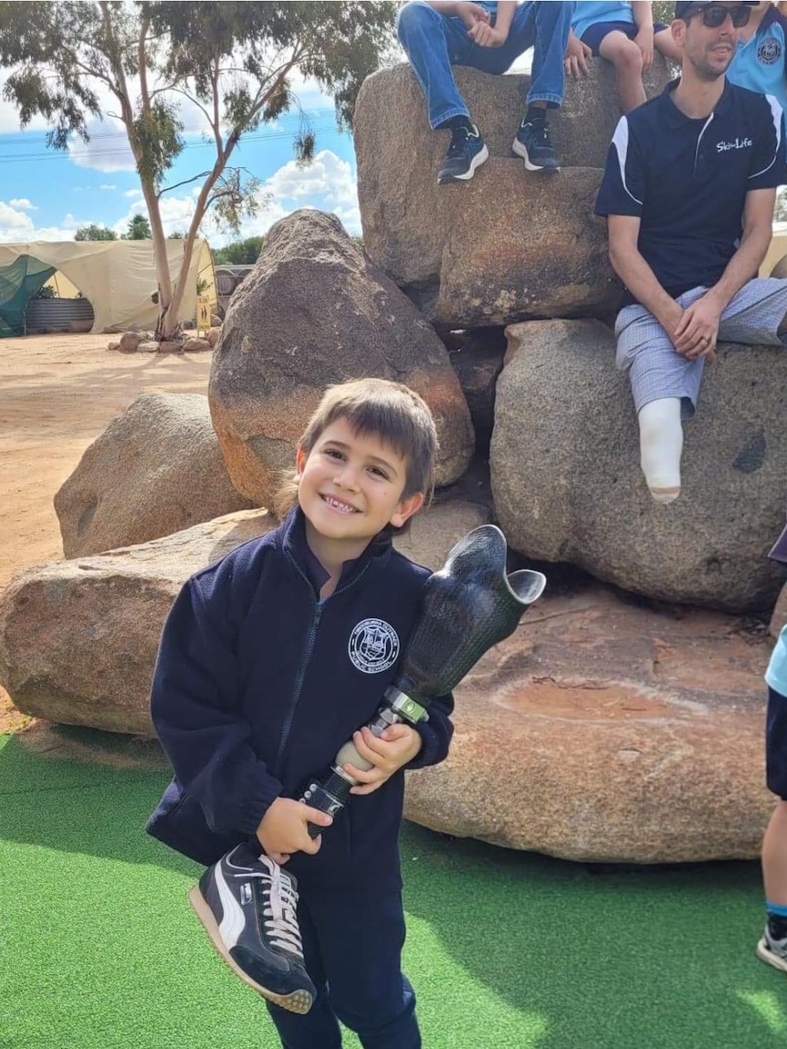 A boy with brown hair smiles while standing on a playground holding a prosthetic leg limb, Ausnew Home Care, NDIS registered provider, disability