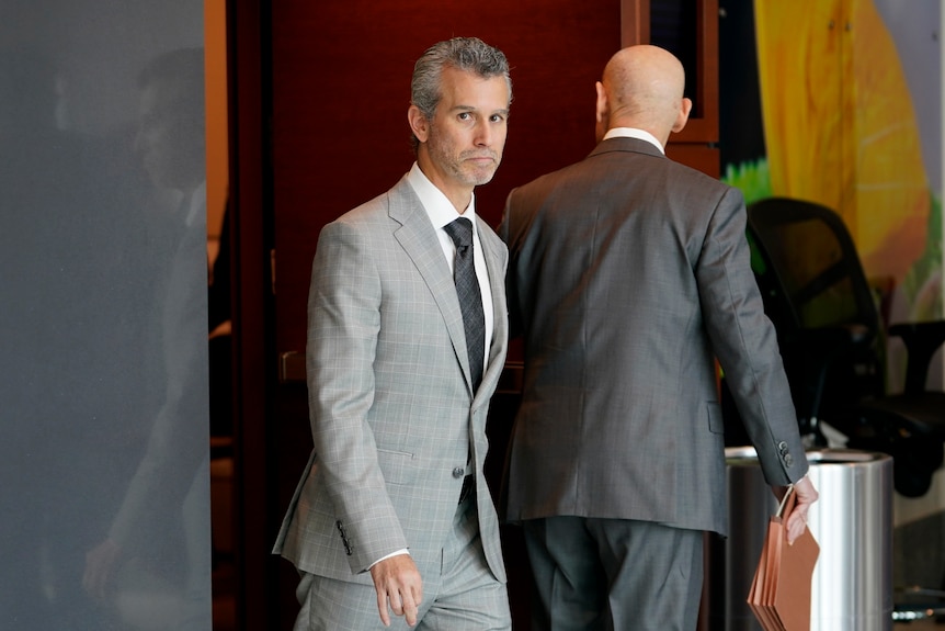 A man in a grey suit walks through the doors of a courtroom.