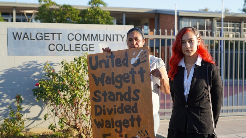 A mother and daughter stand in front of a school entrance with a protest sign