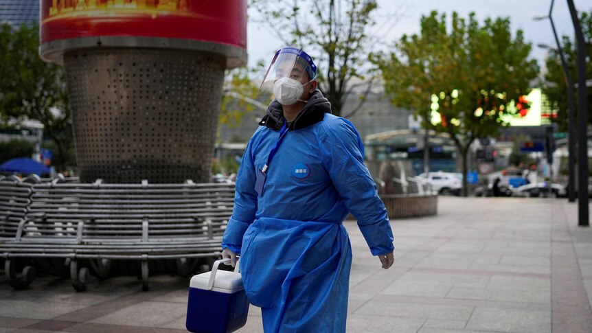 A man in blue protective clothing walks along a street with a small esky in his hand. 
