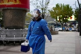 A man in blue protective clothing walks along a street with a small esky in his hand. 