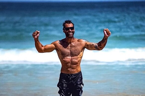 A shirtless man in sunglasses and boardshorts walks out of the surf flexing his biceps