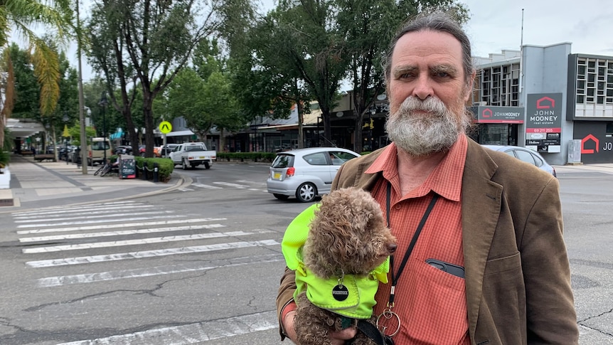 A bearded man wearing an orange shirt and holding a small dog in a raincoat.