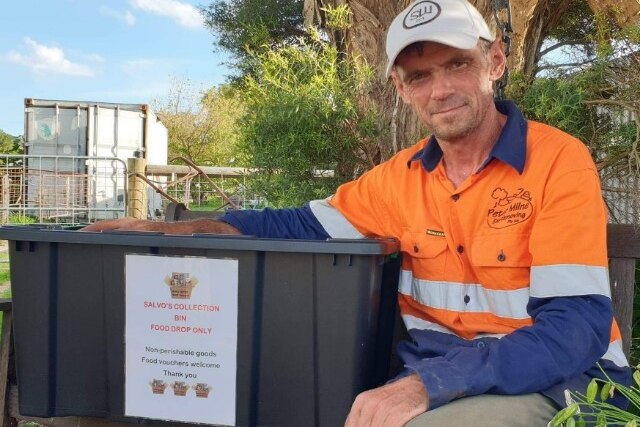 A working man sits with his arm around a crate, he wears a baseball cap and high vis working clothes