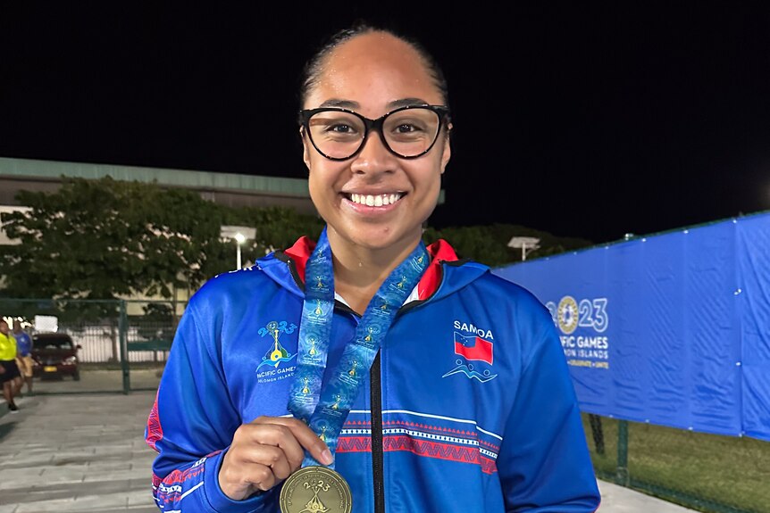 A woman wearing glasses smiles at the camera as she holds up a gold medal.