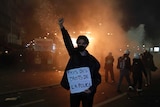 A woman holds a sign saying 'pays des droits de la police' while raising a fist, with fires behind her.