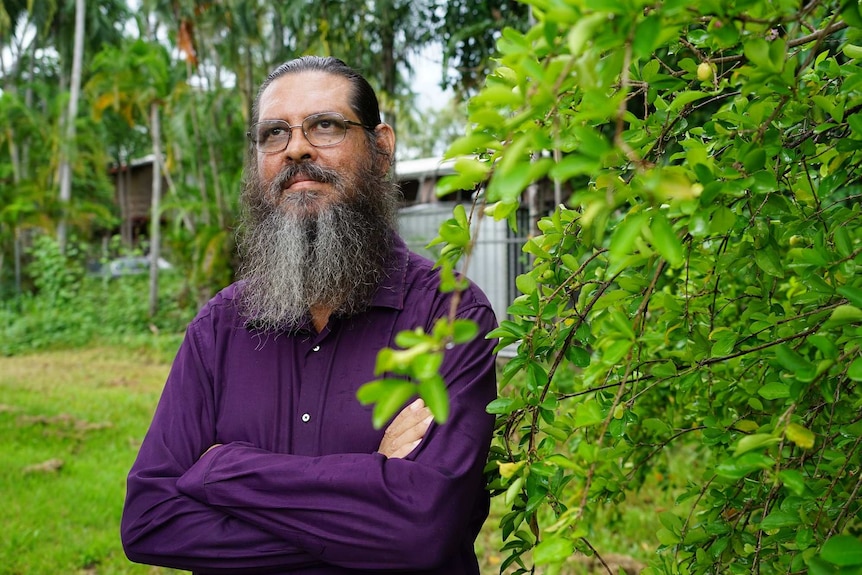 Peter Henwood, with a long beard, purple shirt and glasses, looks out over a green backyard.