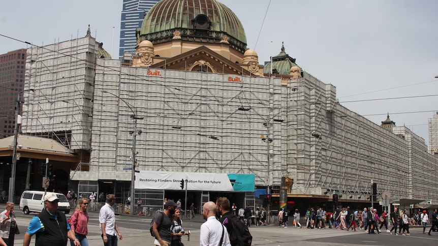 Pedestrians cross in front of a large historic building with a domed roof, the front covered in scaffolding.