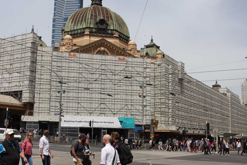 Pedestrians cross in front of a large historic building with a domed roof, the front covered in scaffolding.