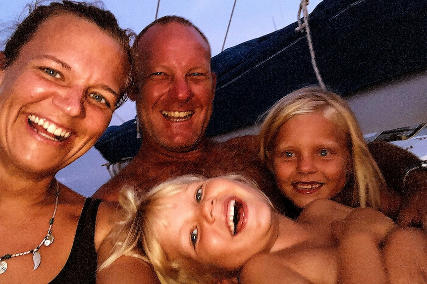 Selfie of family smiling at dawn or dusk with a mainsail cover on a boom behind them.