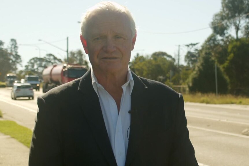 Stephen Caswell stands in suit next to Great Western Highway with trucks behind.