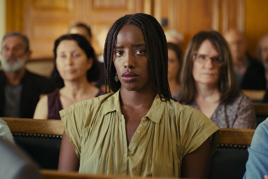 An African American woman sits in a crowded courtroom, with a concerned look in her eyes.