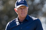 Greg Norman looks down the fairway while he reaches for a club in his bag.