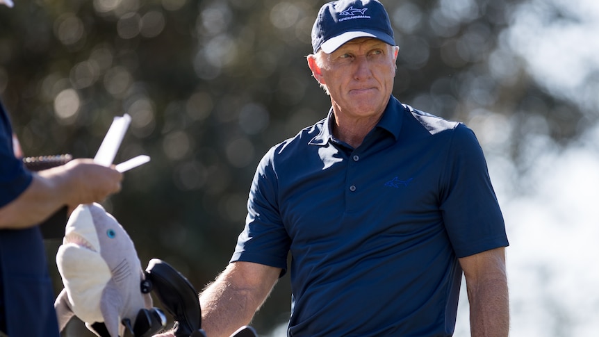 Greg Norman looks down the fairway while he reaches for a club in his bag.