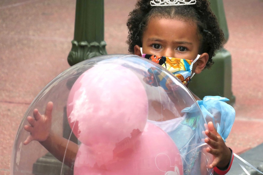 A little girl in a Cinderella costume and mask holds a balloon