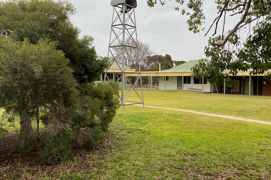 The view of a white wooden building with a green roof and verandah across a lawn and trees.