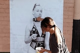 A woman pasting a black and white poster of a girl in lingerie to the wall