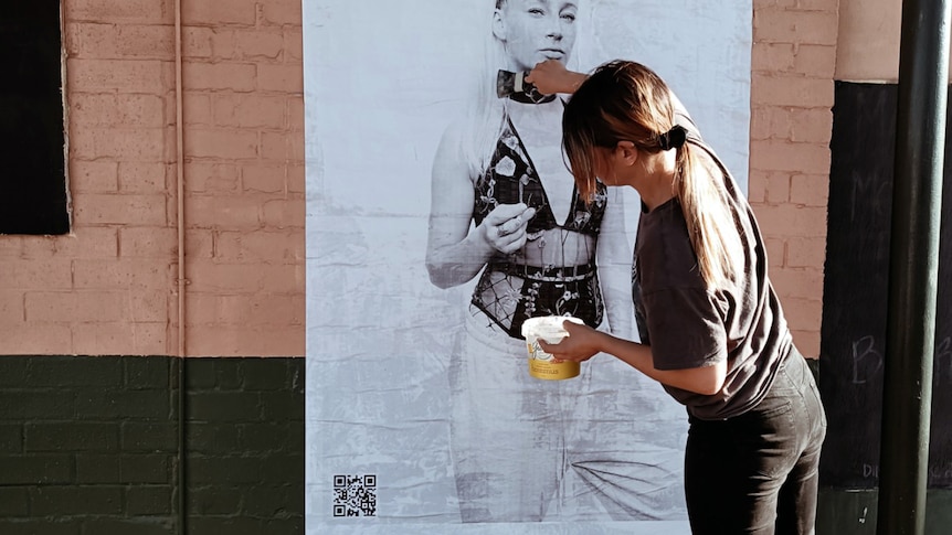 A woman pasting a black and white poster of a girl in lingerie to the wall
