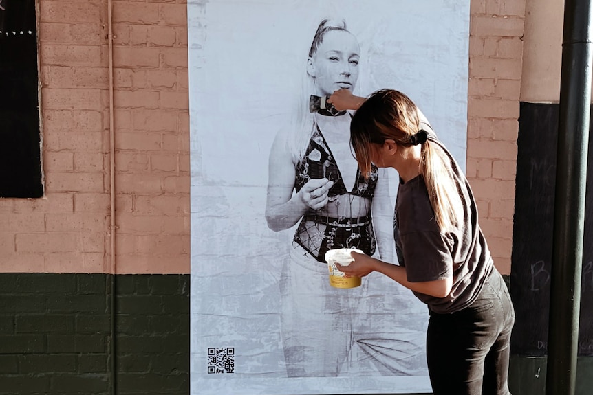 A woman sticks a black and white poster of a girl in lingerie on the wall