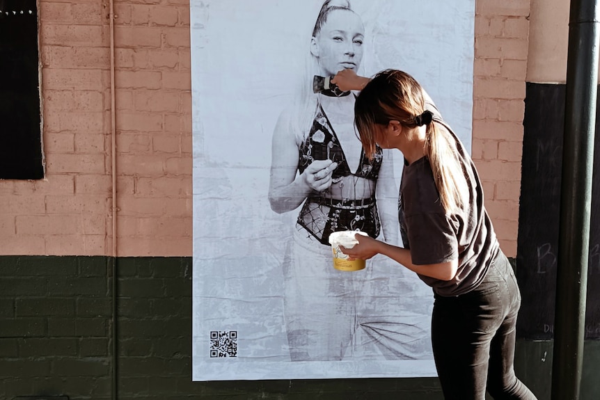 A woman pasting a black and white poster of a girl in lingerie to the wall