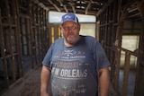 Man standing in a ruined house wearing a blue shirt and hat.