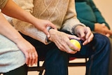 An older person in a sweater squeezes a stress ball while a younger person holds their arm.