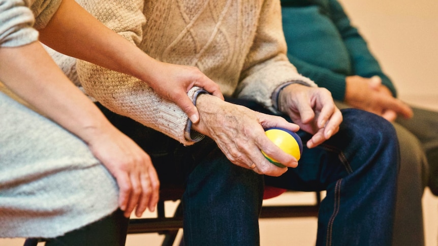 An older person in a sweater squeezes a stress ball while a younger person holds their arm.