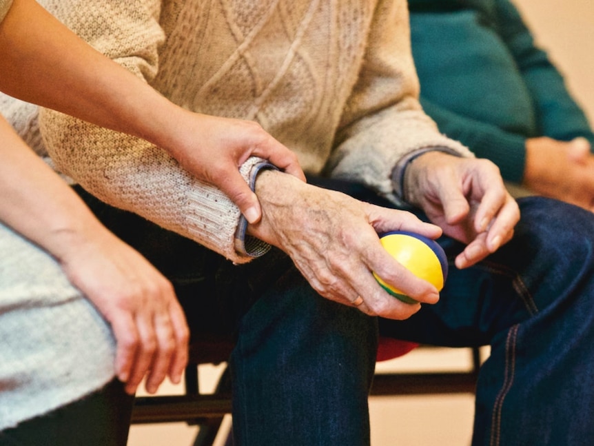 An older person in a sweater squeezes a stress ball while a younger person holds their arm.