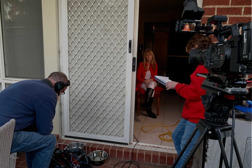 Reporter, sound recordist and camera set up outside open door with woman sitting on chair inside preparing to be interviewed.